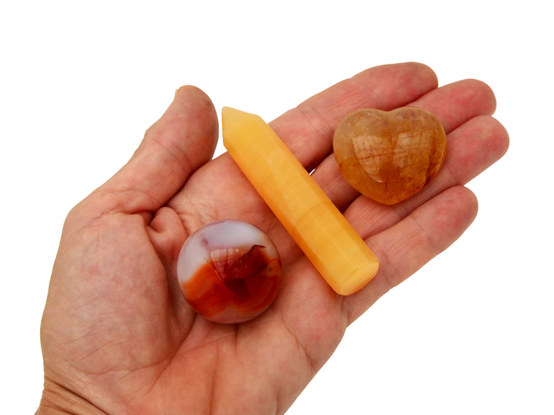 Small faceted orange calcite point, golden healer quartz heart and carnelian sphere on hand with white background