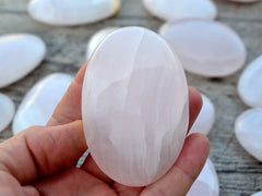 One pink mangano calcite palm stone 80mm on hand with background with some stones on wood table