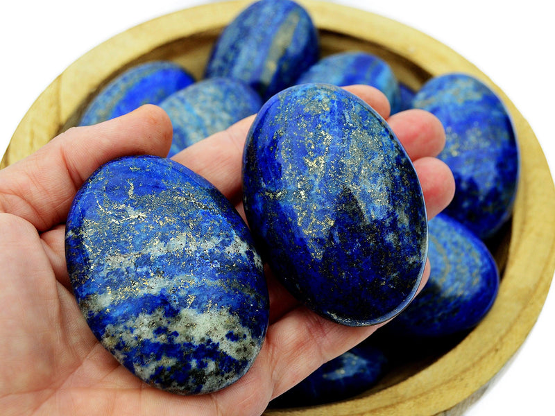 Two lapis lazuli palm stone crystals on hand with background with some stones inside a wood bowl