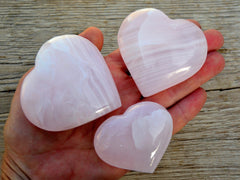 Three mangano calcite heart crystals 50mm-65mm on hand with background with wood 