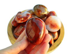 Onre red carnelian tumbled stone on hand with background with some stones inside a wood bowl on white
