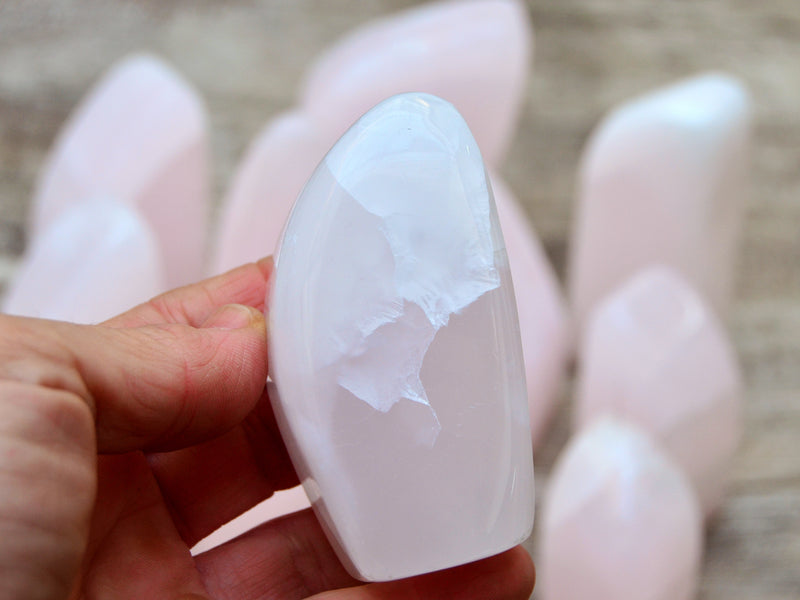 One pink mangano calcite free form 70mm on hand with background with some specimens on wood table