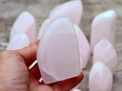 One pink mangano calcite free form 75mm on hand with background with some specimens on wood table