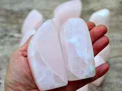 Two pink mangano calcite free form minerals 100mm on hand with background with some stones on wood table