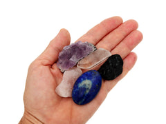 Raw black tourmaline crystal, druzy purple amethyst, raw rose quartz, clear quartz point and lapis lazuli palm stone on hand with white background