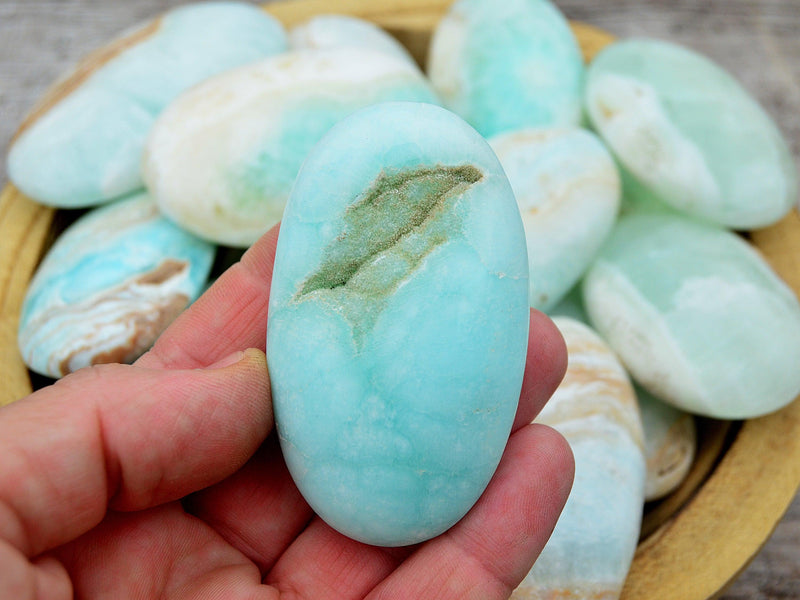 One blue caribbean calcite palm stone 55mm on hand with background with some crystals inside a wood bowl on wood table