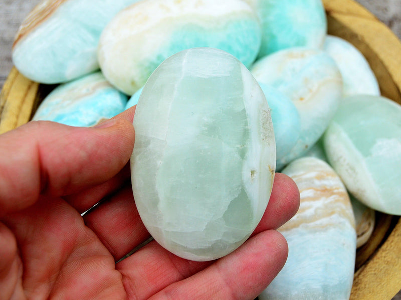 One blue caribbean calcite palm stone 60mm on hand with background with some crystals inside a wood bowl on wood table