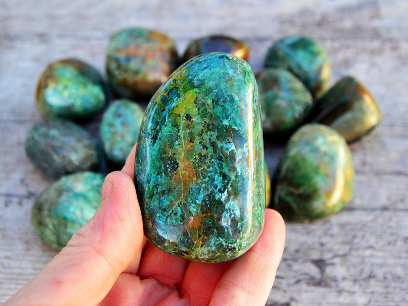 One large green chrysocolla tumbled crystal on hand with background with some minerals on wood table