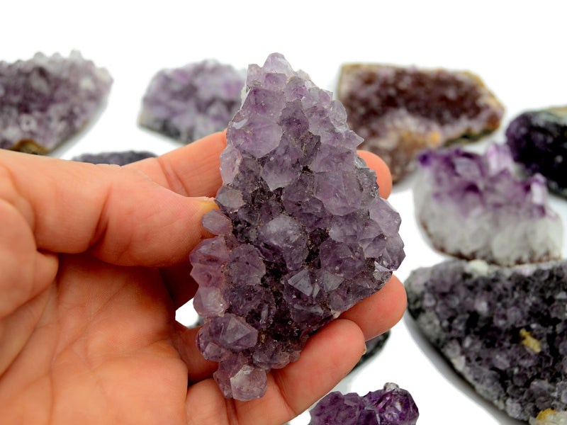Hand holding a purple druzy amethyst crystal with more crystals in the background on a white surface