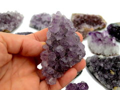 Hand holding a purple druzy amethyst crystal with more crystals in the background on a white surface