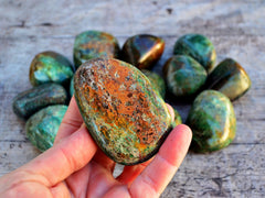 One chunky green and copper chrysocolla tumbled crystal on hand with background with some stones on wood table