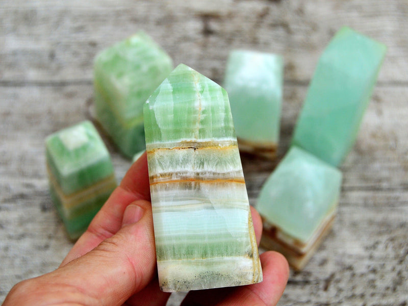 One banded green pistachio calcite tower 60mm on hand with background with some obelisks on wood table