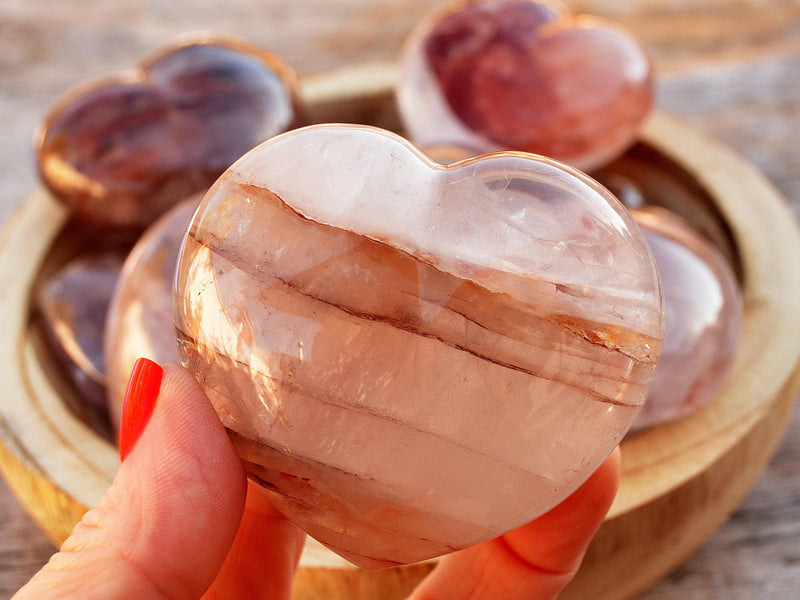 One large hematoid quartz heart stone 67mm on hand with background with some hearts inside a wood bowl