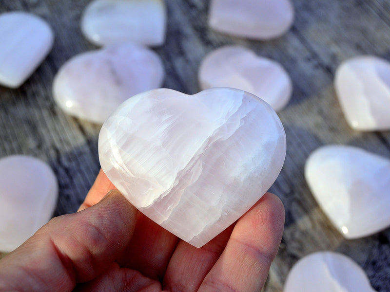One pink mangano calcite crystal heart 50mm on hand with background with several stones on wood table