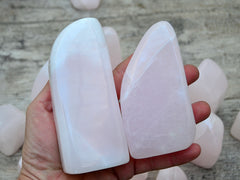 Two pink mangano calcite free form crystals 100mm on hand with background with some specimens on wood table