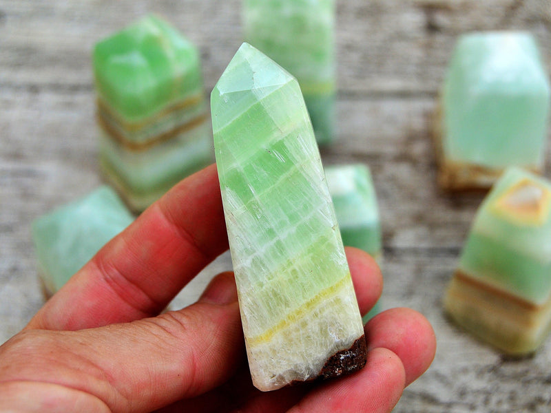 One banded green pistachio calcite obelisk 65mm on hand with background with some obelisks on wood table