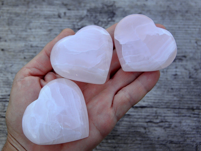 Three pink mangano calcite hearts 55mm-60mm on hand with wood background