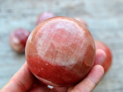 One rose calcite sphere crystal 65mm on hand with background with some balls on wood table