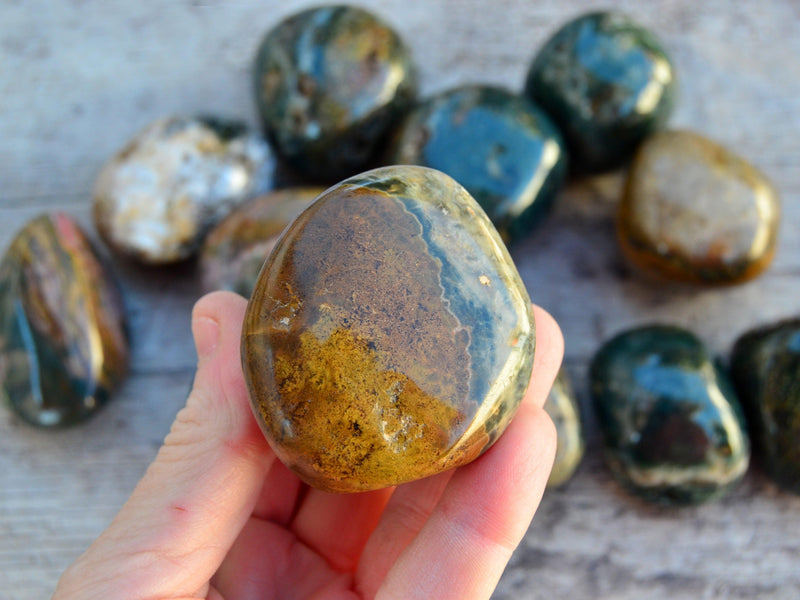 One smustard yellow and bown ocean jasper tumbled stone on hand with background with some minerals on wood table