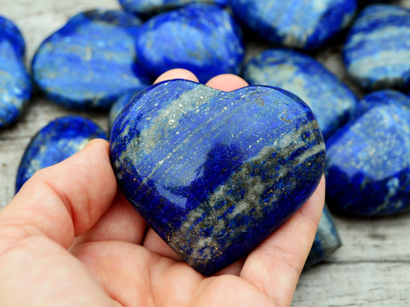 One lapis lazuli heart crystal 55mm on hand with background with some crystals on wood table
