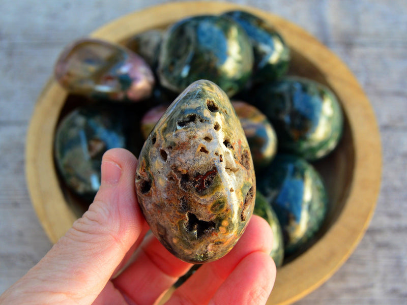 One green ocean jasper tumbled stone on hand with background with some minerals inside a wood bowl on wood table