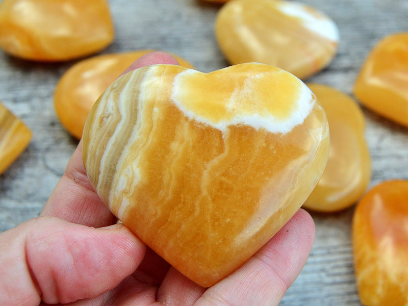 One orange calcite heart shapped stone 60mm on hand with background with some stones on wood table