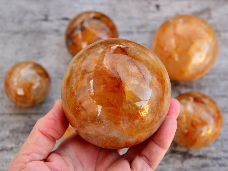 One golden healer quartz sphere 75mm on hand with background with some hematoid balls on wood table
