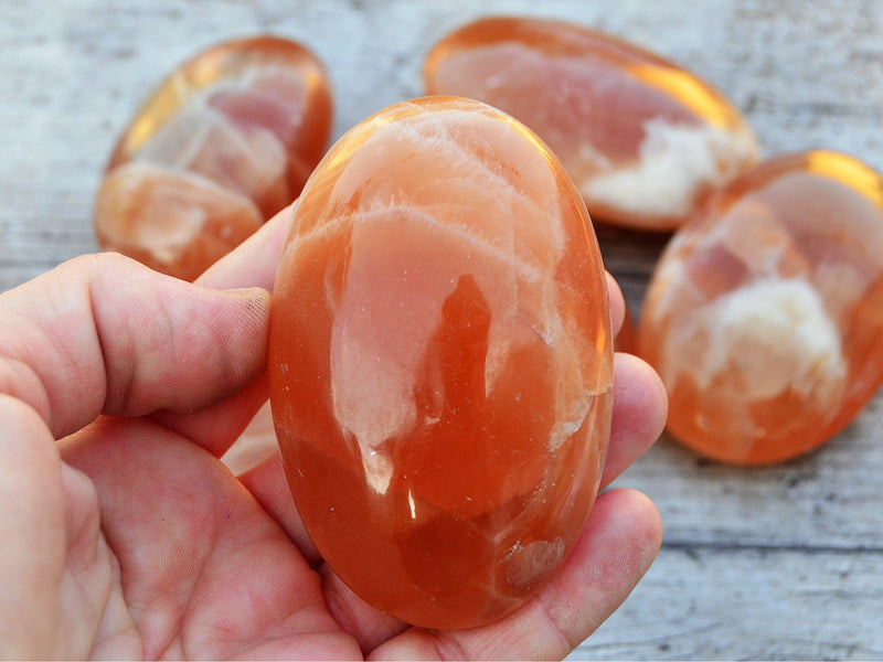 One extra large honey calcite palm stone crystal 85mm on hand with background with some stones on wood table
