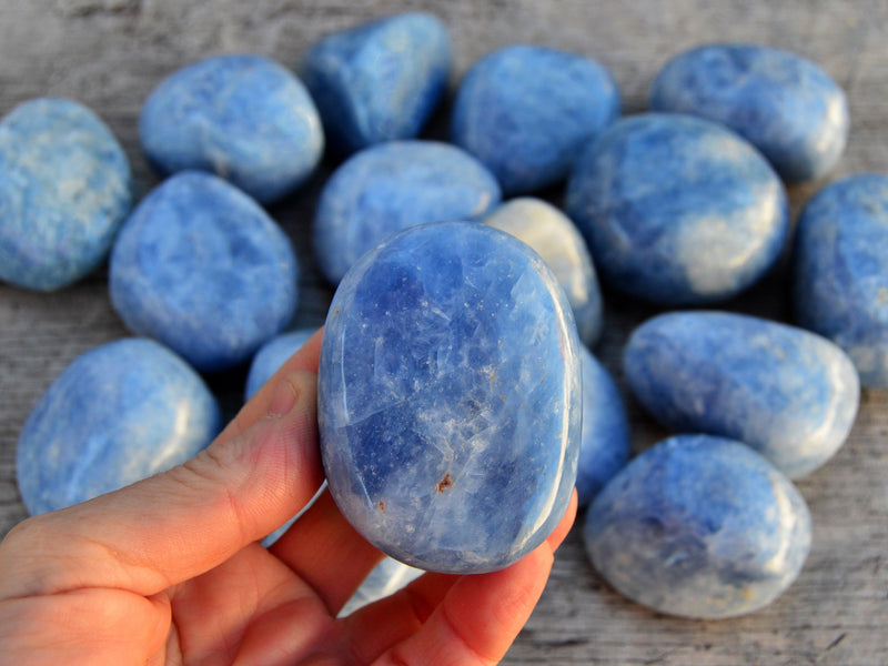 One chunky blue calcite tumbled crystal on hand with background with some minerals on wood table