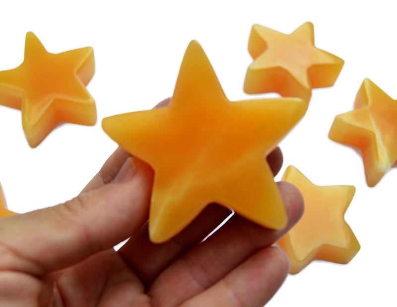 One orange calcite star carving gemstone 60mm on hand with background with some stones on white