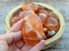 One orange calcite plam stone 55mm on hand with background with several crystals inside a wood bowl on wood table
