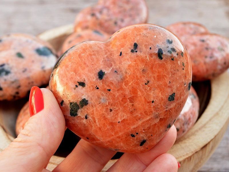 One large orange calcite crystal heart 65mm on hand with background with some hearts inside a wood bowl