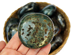 One green sea jasper palm stone 55mm on hand with background with some stones inside a straw basket