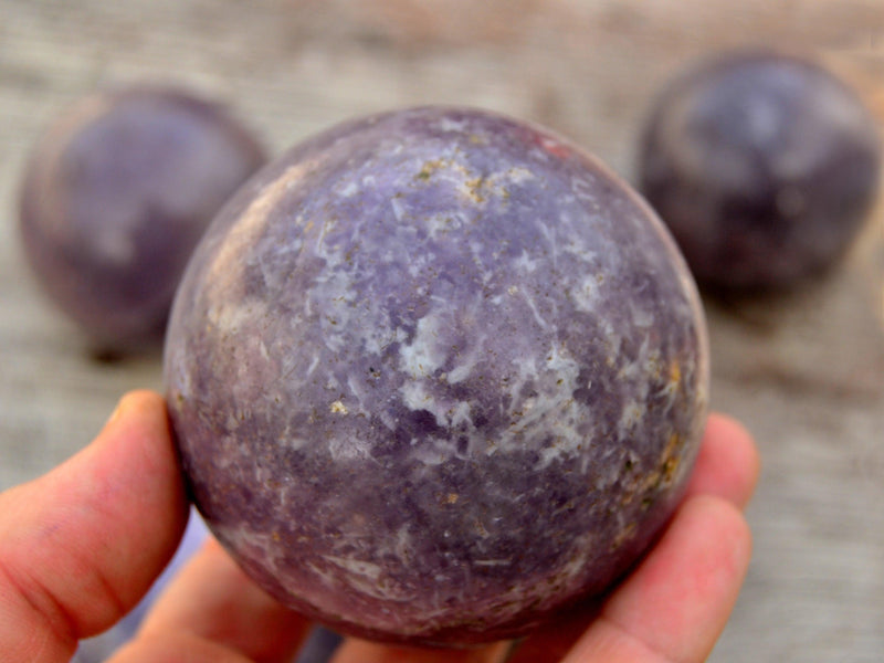 Large lepidolite crystal sphere 75mm on hand with background with some balls on wood table