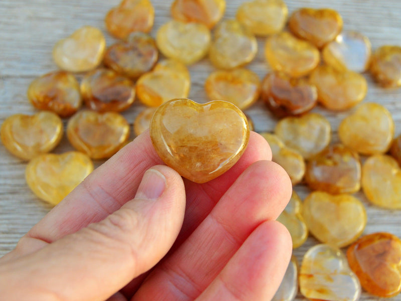 One yellow hematoid quartz crystal heart 30mm on hand with background with several hearts on wood table