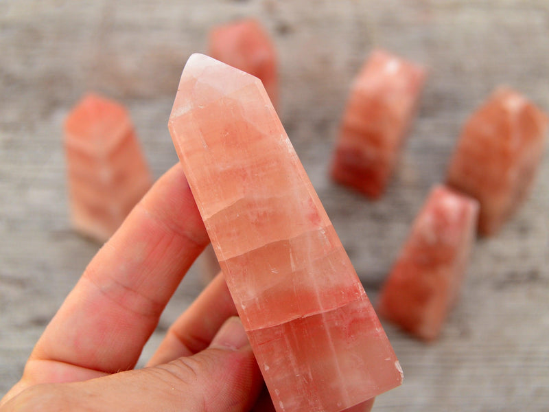 One rose calcite obelisk crystal 80mm on hand with background with some crystals on wood table
