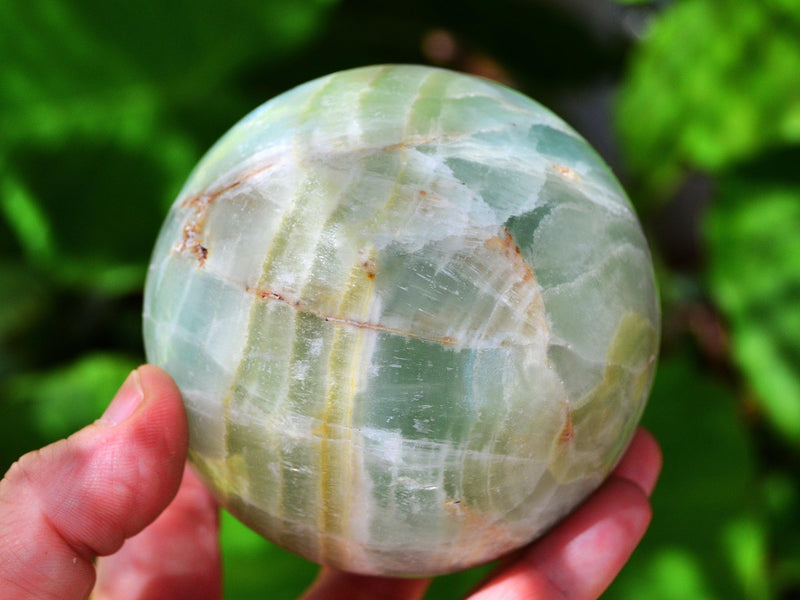 Large green pistachio calcite sphere 90mm on hand with background with green plants