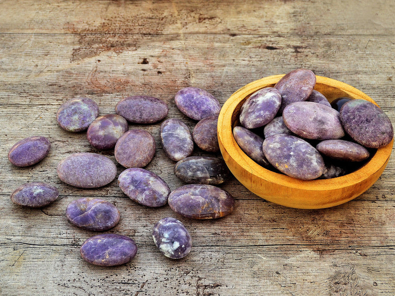 Several purple lepidolite palm stones inside a wood bowl with background with some palm stones on wood table