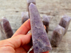 One large lepidolite tower 115mm on hand with background with some obelisks on wood table