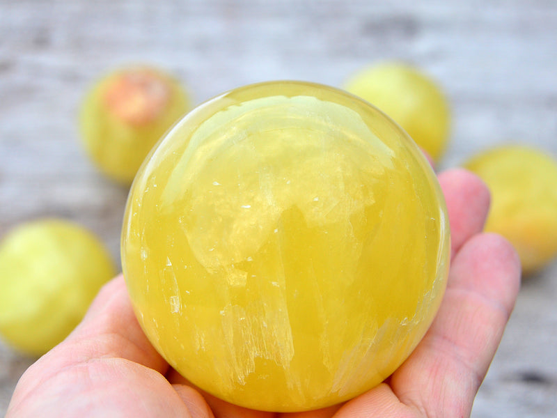 One lemon yellow calcite sphere crystal 60mm on hand with background with some stones on wood table