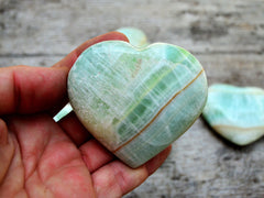 One green banded pistachio calcite heart 55mm on hand with background with some stones on wood table