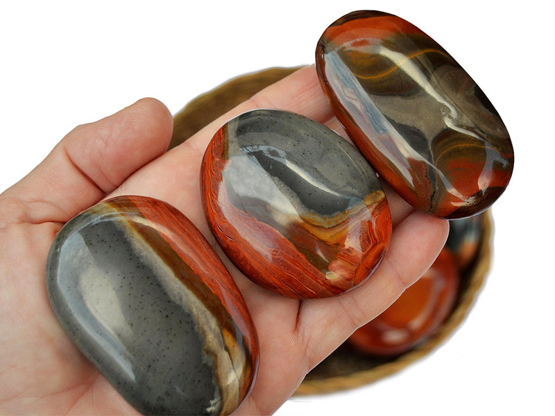 Three multicolor polychrome jasper palm stones 40mm - 70mm on hand with background with some crystals inside a basket