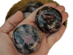 Two rhodonite palm stones on hand with background with some stones inside a basket on white