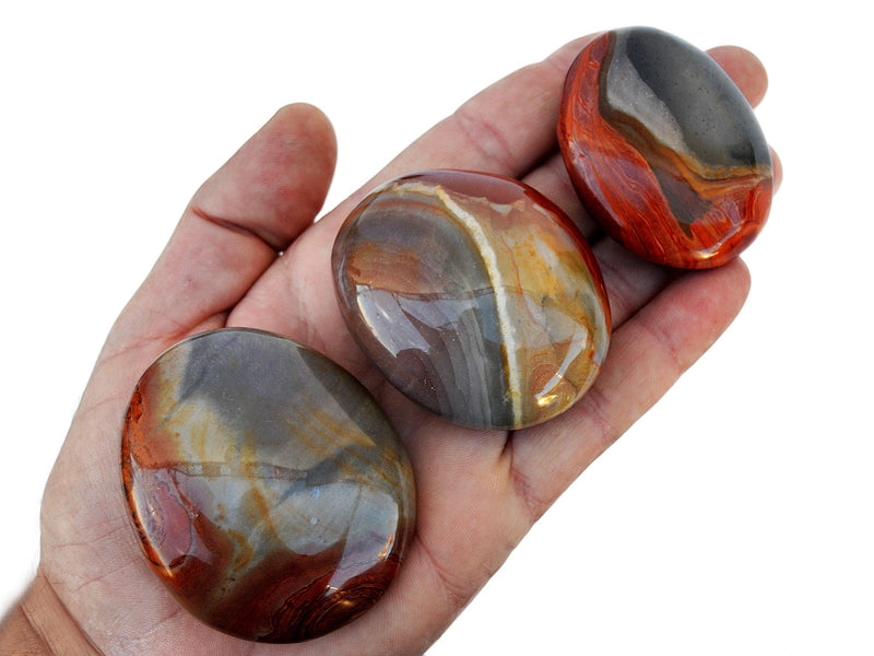 Three multicolor desert jasper palm stones 40mm - 70mm on hand with background with some crystals on white