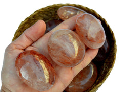 Three hematoid quartz palm stones 40mm-70mm on hand with background with some gemstones inside a straw basket on white