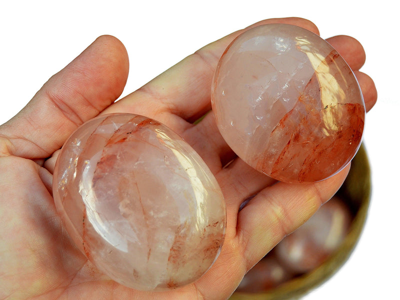 Two hematoid quartz palm stones 60mm-70mm on hand with background with some gemstones inside a straw basket on white