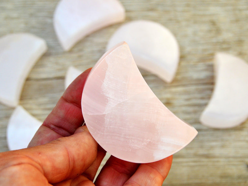 One pink mangano calcite shhaped moon crystal 60mm on hand with background with some moons on wood table