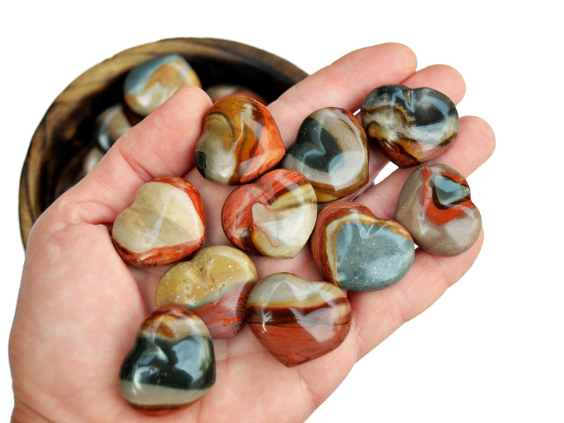 Ten small polychrome jasper crystal hearts on hand with background with some crystals inside a wood bowl