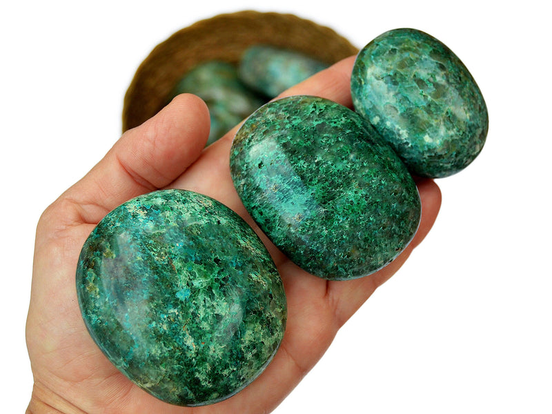 Three chrysocolla palm stones 50mm-70mm on hand with background with some crystals inside a basket