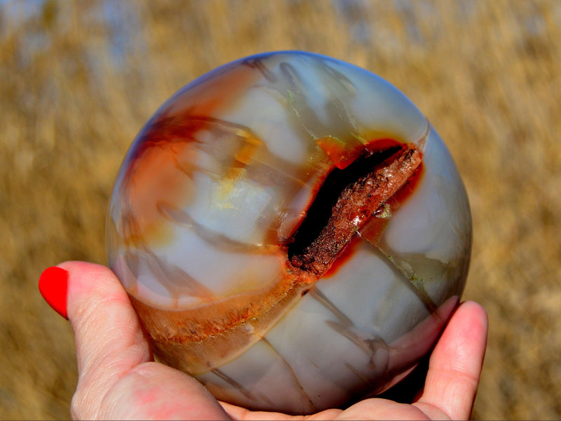 One large carnelian crystal sphere 100mm on hand with straw landscape background